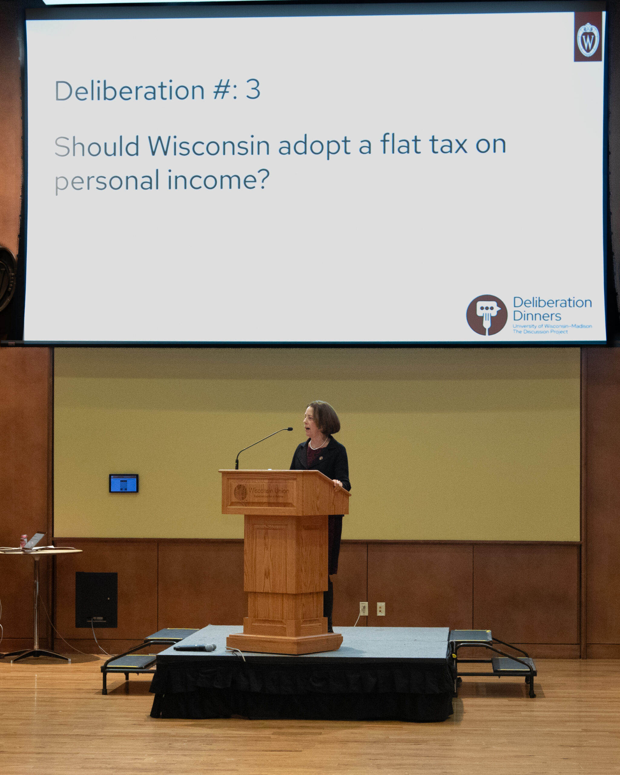 Dean Hess at a podium on stage with a projected slide that says Deliberation number 3: Should Wisconsin adopt a flat tax on personal income?