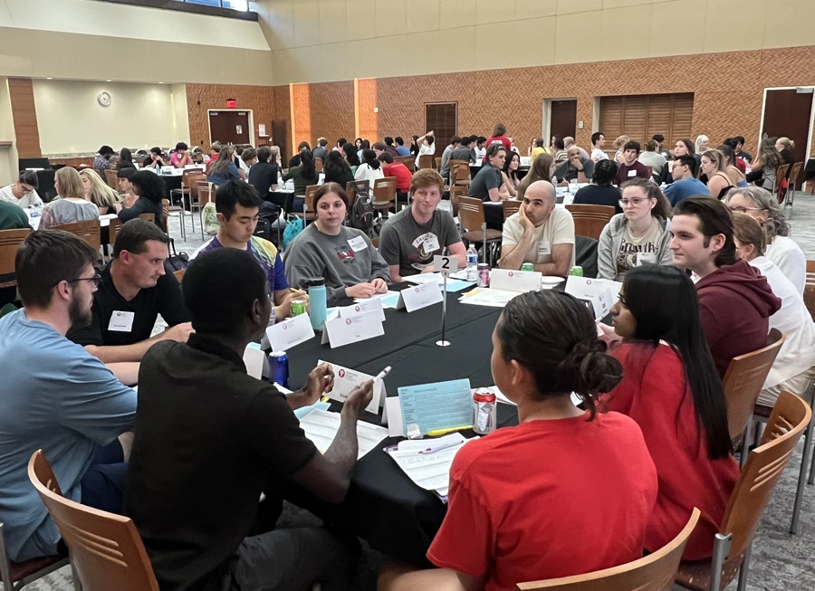 Group of students sitting at a table in a large room with other groups at tables.