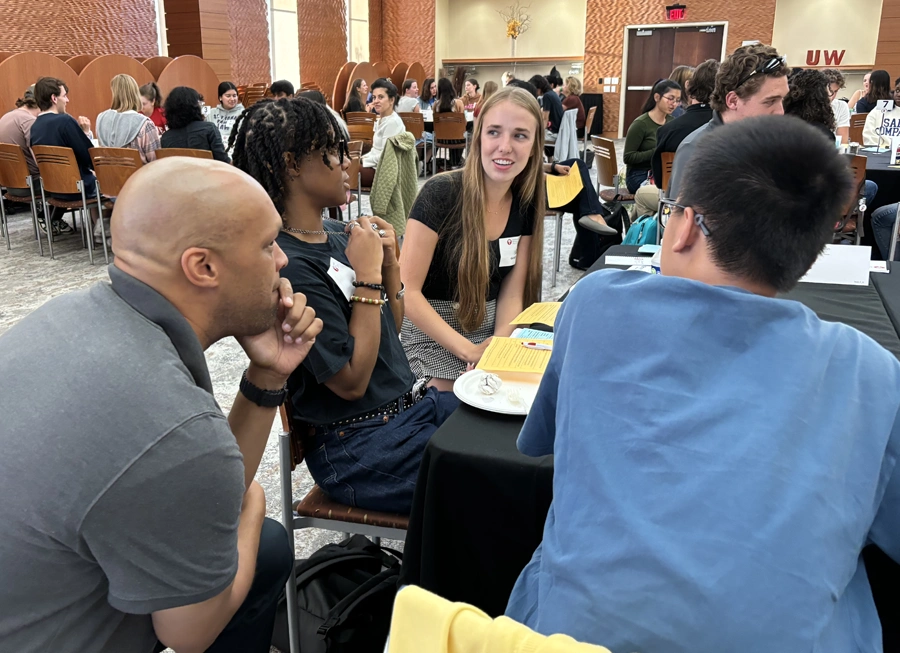 Two students are having a conversation while sitting at a table. A facilitator is sitting next to them and is leaning forward.