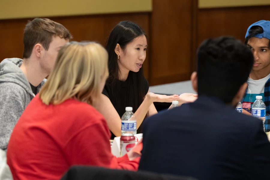 A student is talking at a table and a few people are sitting next to them.