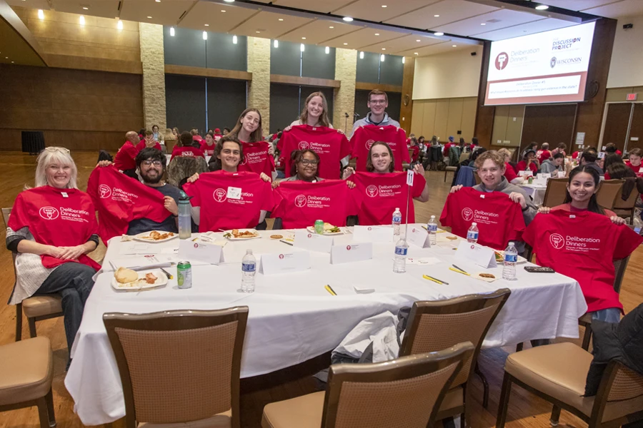 A group of students with a facilitator at a table and everyone is holding up a red Deliberation Dinners t-shirt.
