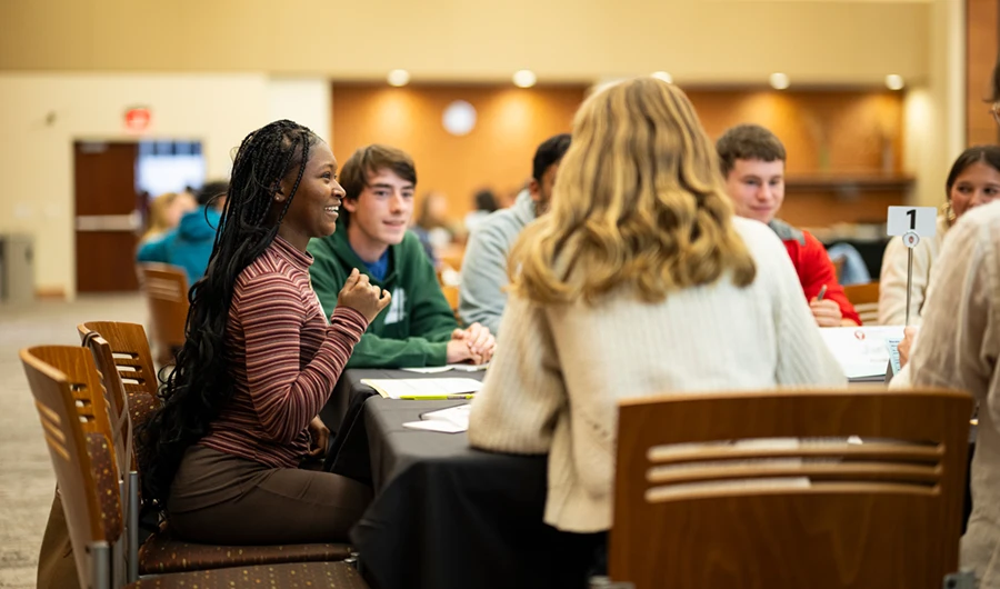 Side view of a student sitting at a table with others. The student is leaning forward and smiling.